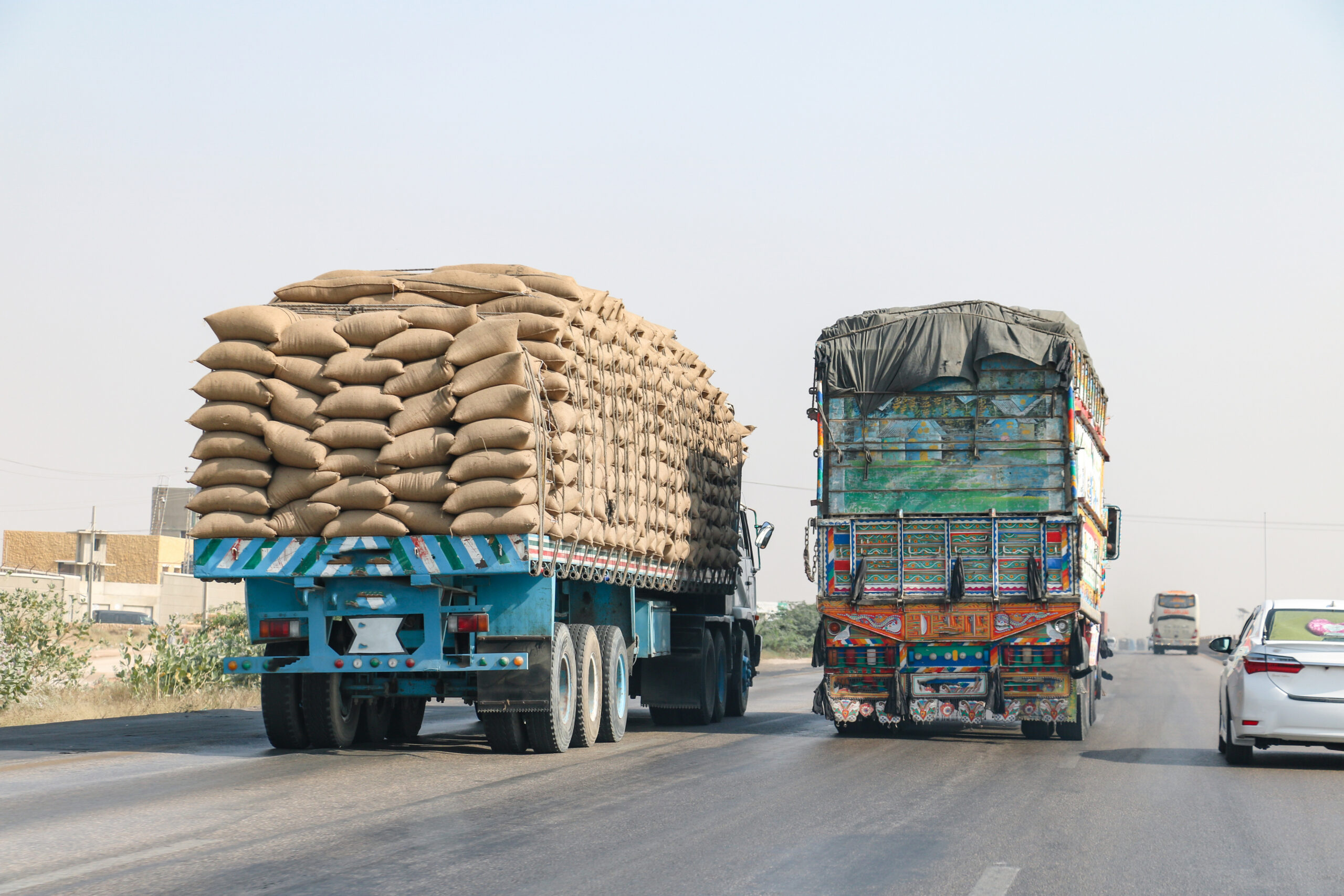 Two heavily loaded commercial trucks travel on a Pennsylvania highway, with stacked burlap sacks and a covered cargo bed beside passing traffic.