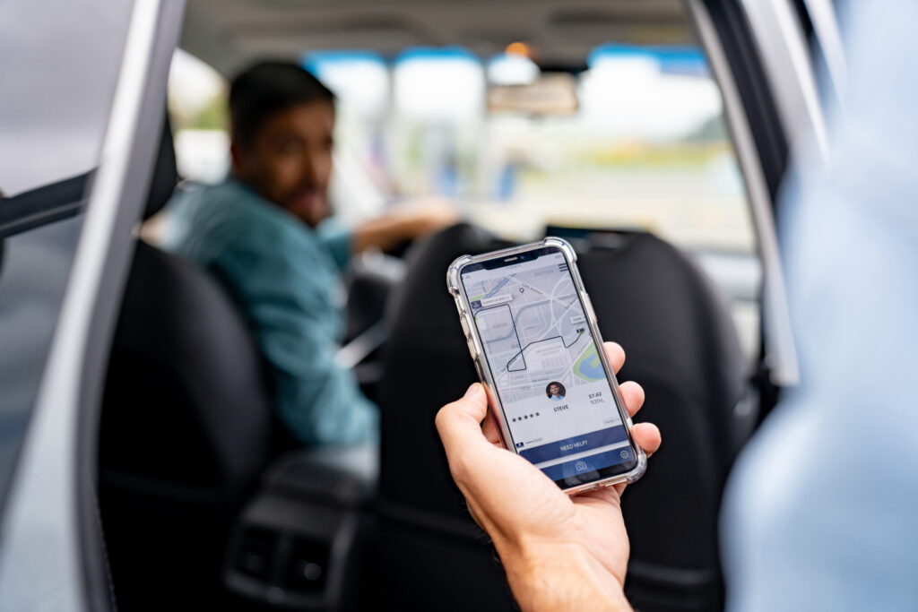 A passenger holds a rideshare app phone in the back seat while a driver sits ahead, reflecting Uber and Lyft travel in Philadelphia traffic.