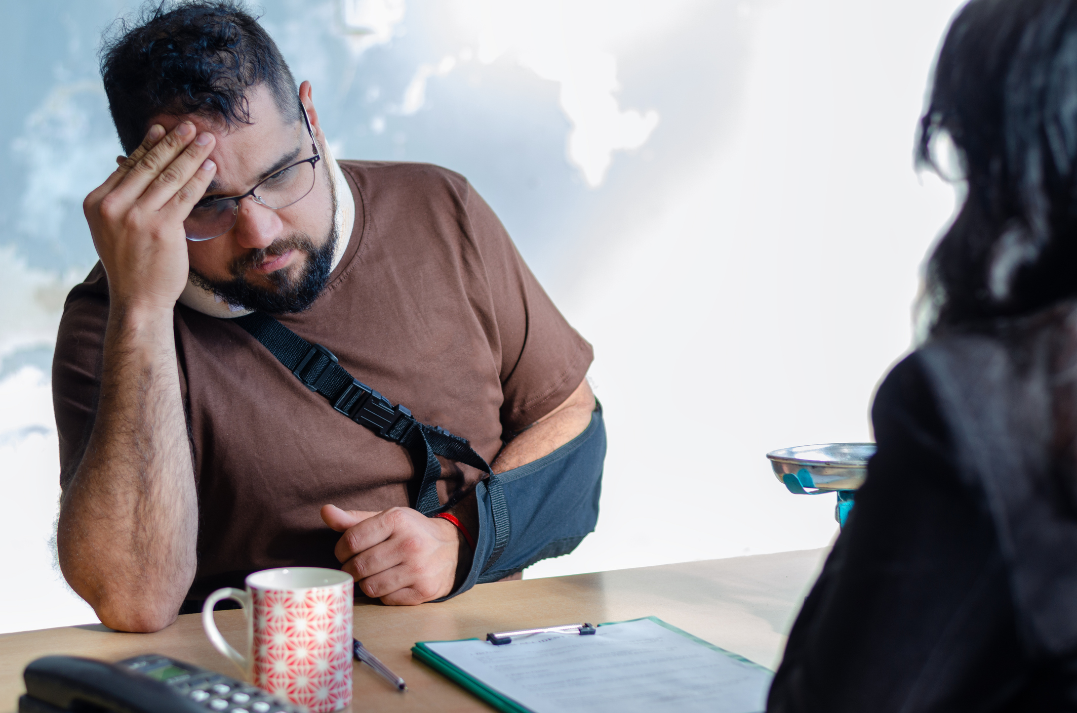 An injured man with a broken arm sits across from a clinician, holding his forehead during a consultation about pain and suffering after a crash.