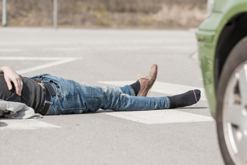 A pedestrian lies injured in the road after a hit-and-run in Philadelphia, with a green car nearby and the driver's foot still in the scene on the asphalt.